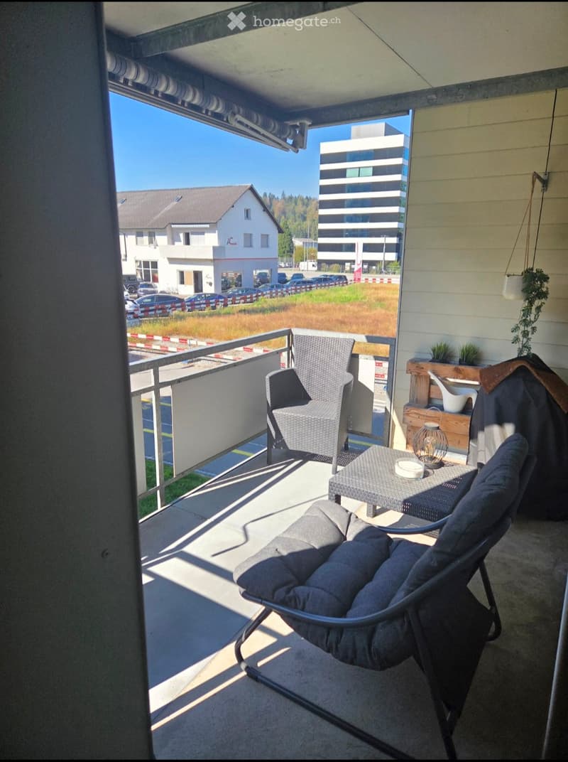 Balcony with a grey wicker chair, a small table, and a hanging lamp, overlooking a parking lot and buildings.