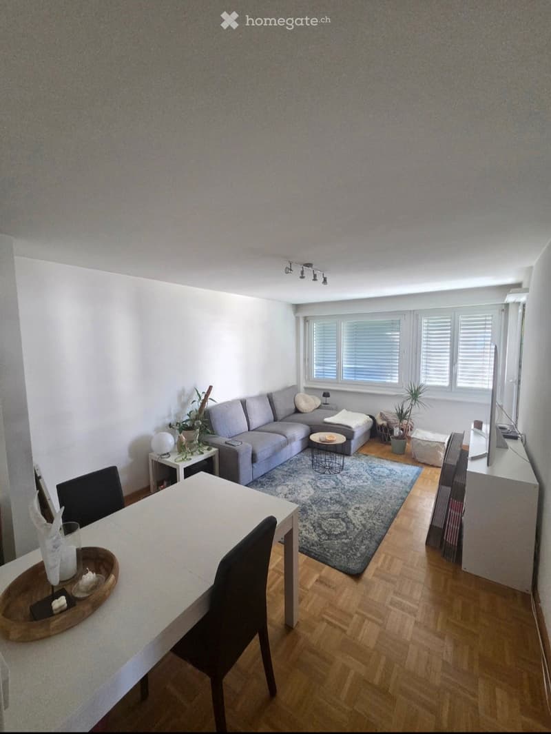 Modern living and dining area with a grey sectional sofa, coffee table, side table, dining table with chairs, and a large window with blinds.