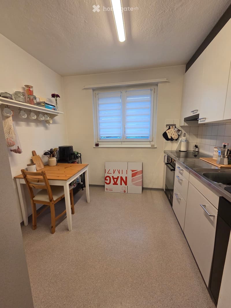 Compact kitchen with white cabinets, a wooden dining table with a chair, a window with blinds, and various kitchen appliances and utensils.