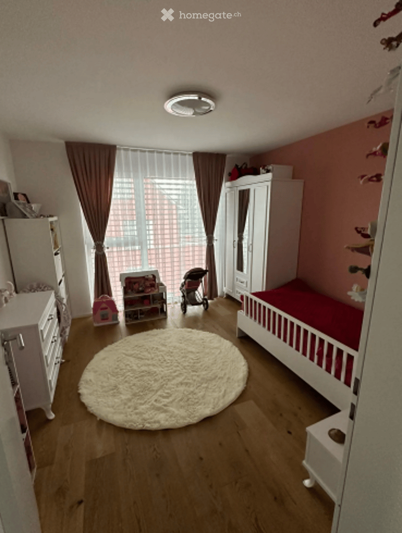 A well-lit children's bedroom with a wooden floor, a white wardrobe, a dresser, a round white rug, a bed with red bedding, and a toy box. A baby stroller is visible near the window with brown curtains.