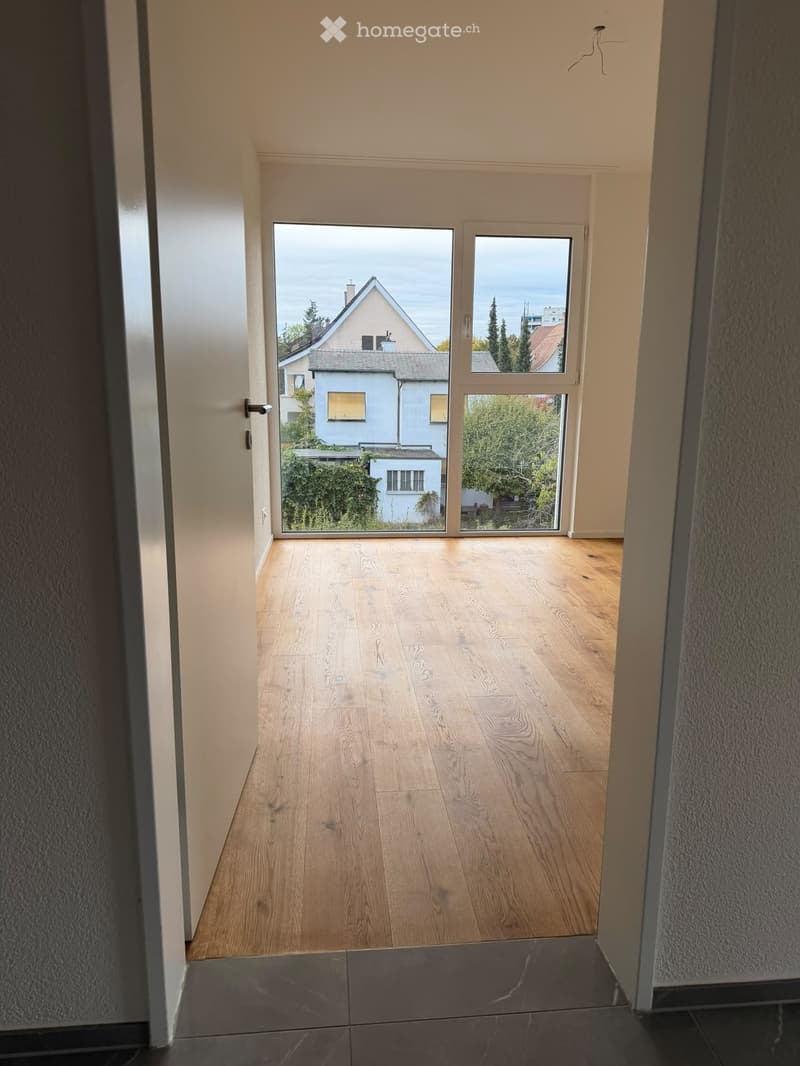 Empty hallway with wooden flooring, large windows, and a view of neighboring houses and greenery.