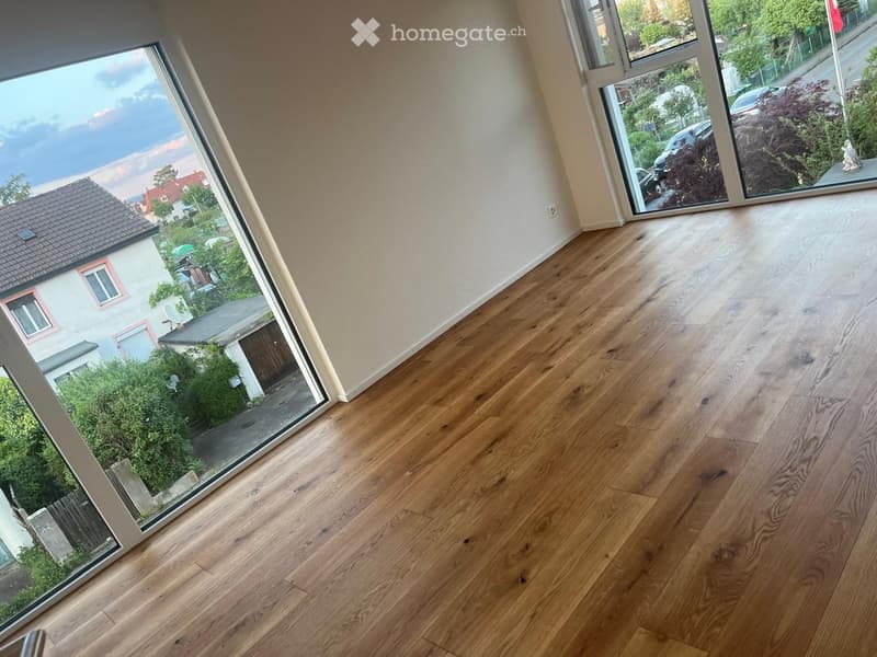 Empty room with wooden flooring and large windows offering views of the street and neighboring houses.