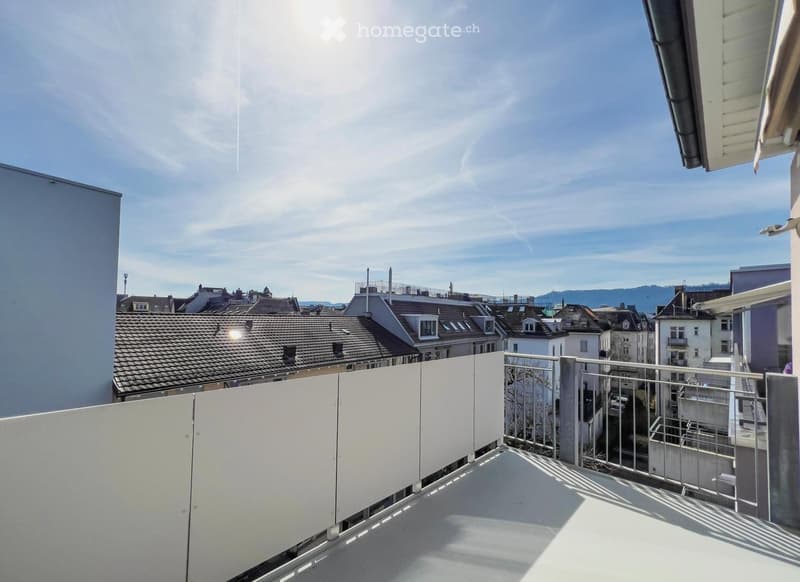 Balcony with white walls and metal railing, overlooking a cityscape with rooftops and distant mountains under a clear blue sky.