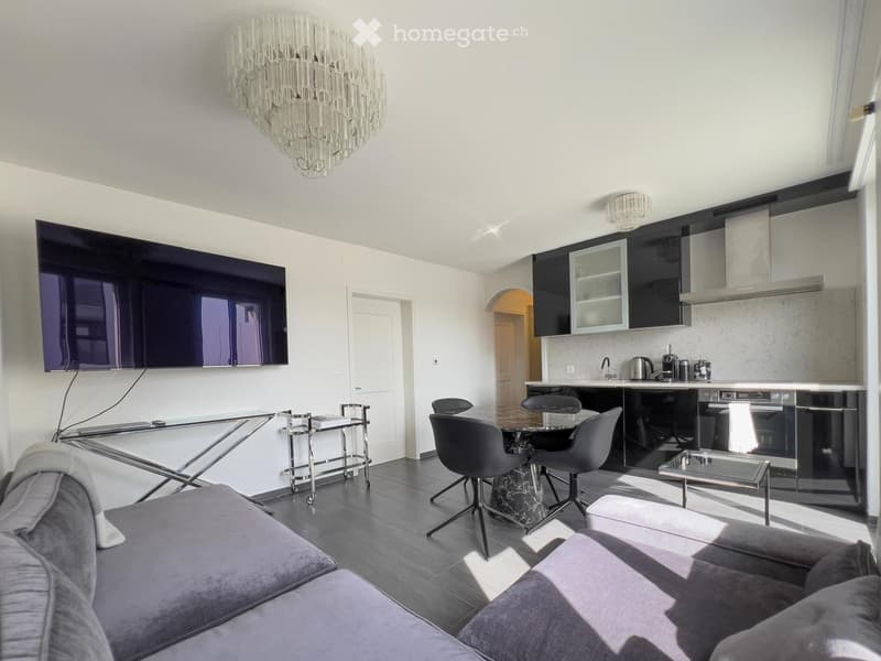 Modern open-plan living and dining area with a grey sofa, coffee table, dining table with chairs, and a sleek kitchen area with cabinets and appliances.