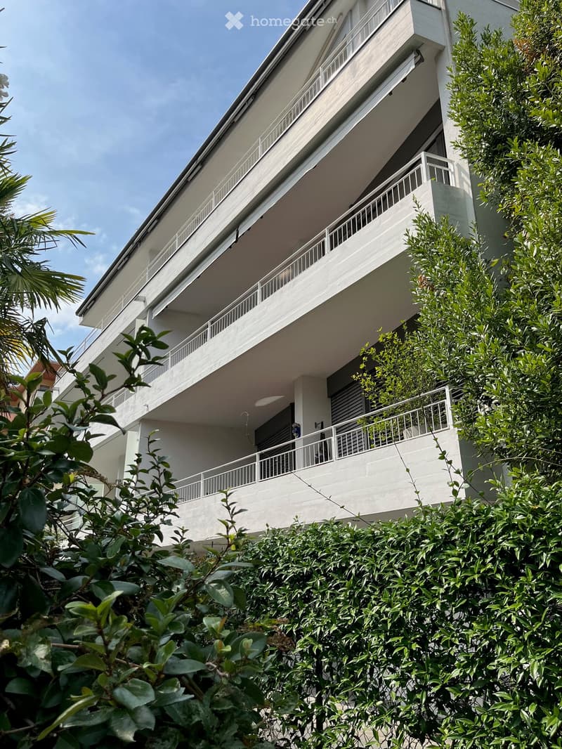 Modern multi-story building with white balconies and railings, surrounded by lush green foliage.