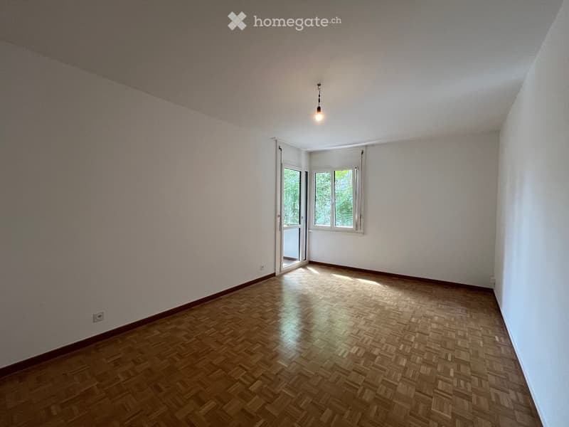 Empty room with wooden parquet flooring, white walls, and a single hanging light fixture. Features a large window and a door leading outside.
