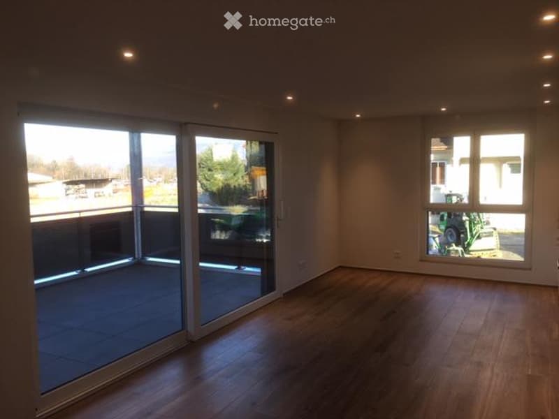 Empty room with wooden flooring, large sliding glass doors leading to a balcony, and a window with a view of the outside area.