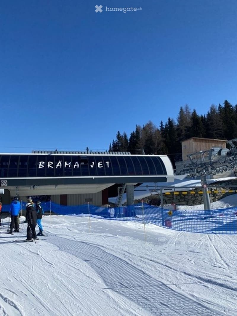 Ski lift station named "BRAMA JET" with a modern, curved structure and a clear blue sky. Skiers are visible on the snow-covered ground.