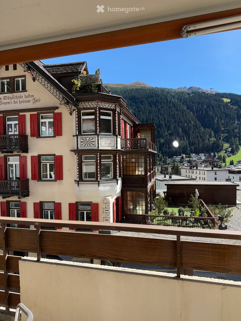 A multi-story building with ornate wooden balconies and red shutters, overlooking a scenic mountain landscape with a town below.