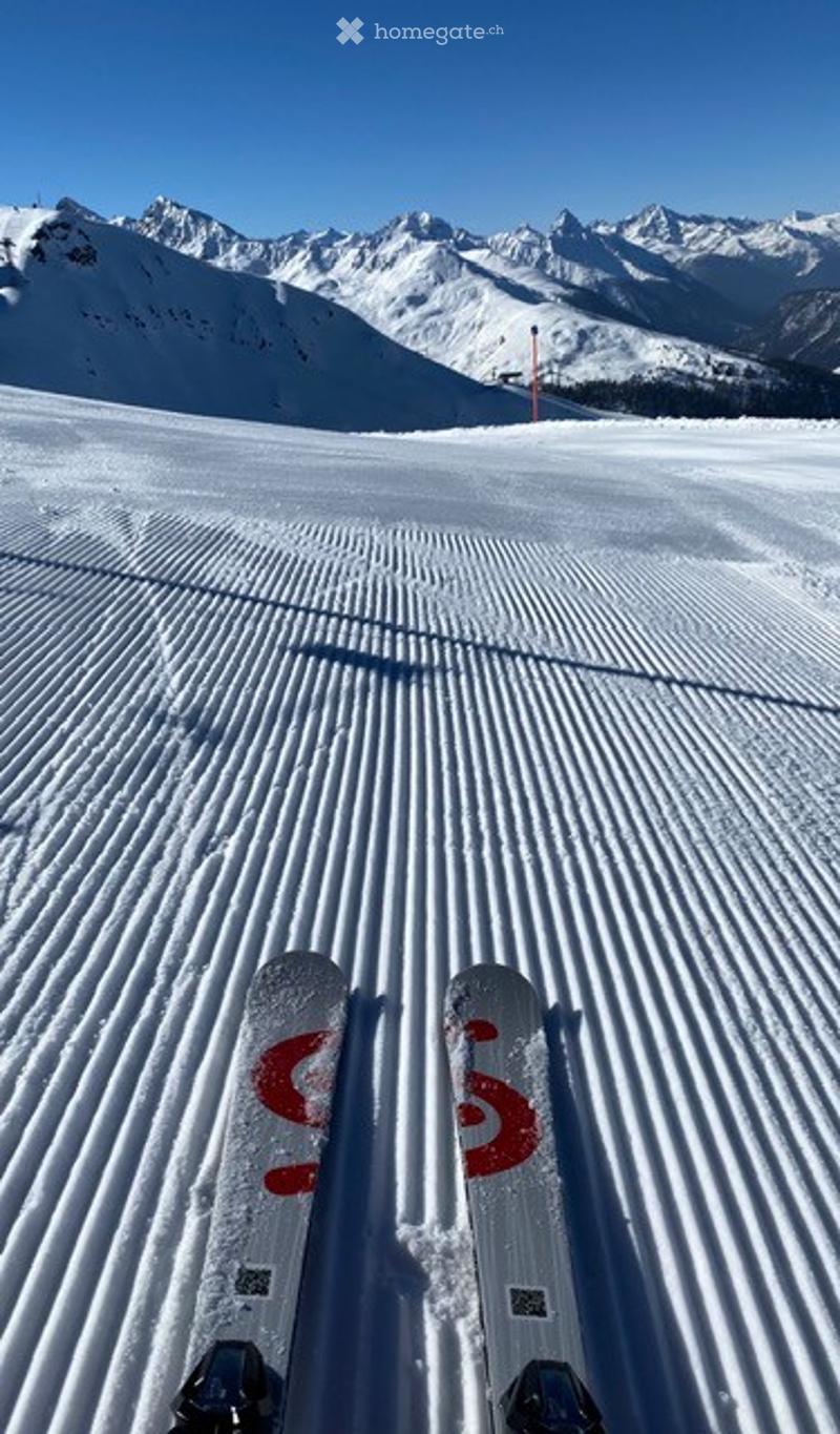 Snow-covered ski slope with groomed tracks, distant snow-capped mountains, and a ski lift pole visible in the background.