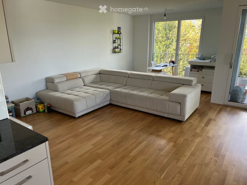 Modern living room with light-colored sectional sofa, wooden flooring, and large windows leading to a balcony. Includes a small table, chairs, and a shelf with decorative items.