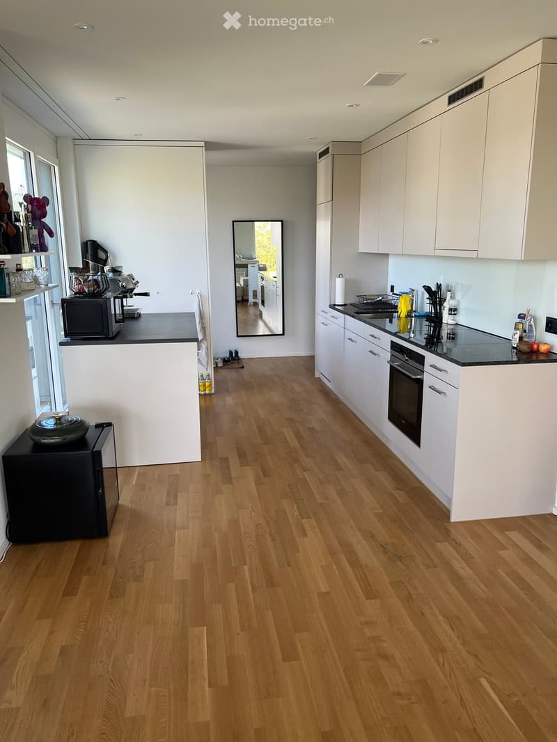 Modern kitchen with white cabinets, wooden flooring, and various appliances including a refrigerator, microwave, coffee maker, and oven. The kitchen features a black countertop, a sink, and a mirror on the wall.