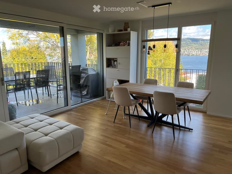 Modern living and dining area with hardwood floors, large windows offering a scenic view, a sofa, a dining table with chairs, and a shelf with decorative items.
