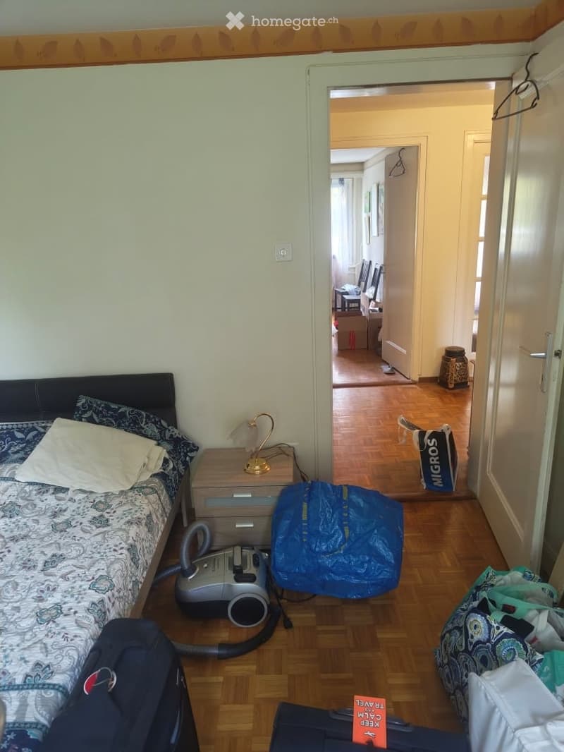 Bedroom with a bed, nightstand, lamp, vacuum cleaner, and various bags and suitcases on the floor. The room has wooden flooring and light-colored walls.