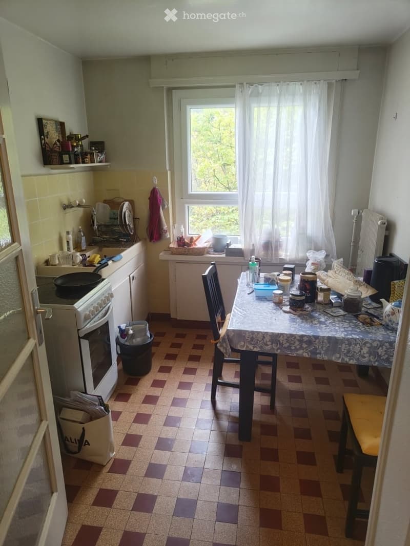 The kitchen features a tiled floor with a mix of brown and purple tiles, a window with white curtains, and a small table with chairs. The kitchen includes an oven, fridge, and various kitchen items on the counter and shelves.
