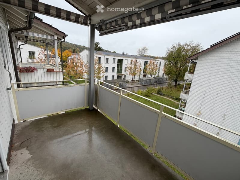 Balcony with concrete floor, metal railing, and awning, overlooking a residential area with buildings and trees.