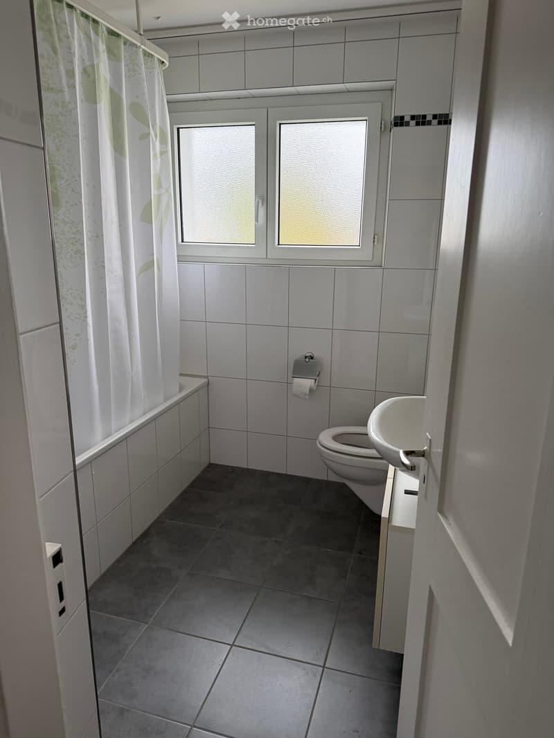 Small bathroom with grey tiled walls and dark grey tiled floor, featuring a toilet, sink, bathtub, and a window with frosted glass panes.