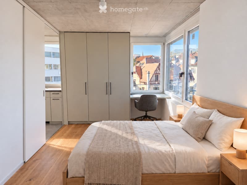 Modern bedroom with wooden flooring, large windows offering a cityscape view, a neatly made bed with pillows and a blanket, a nightstand with a lamp, a desk with a chair, and built-in wardrobes.