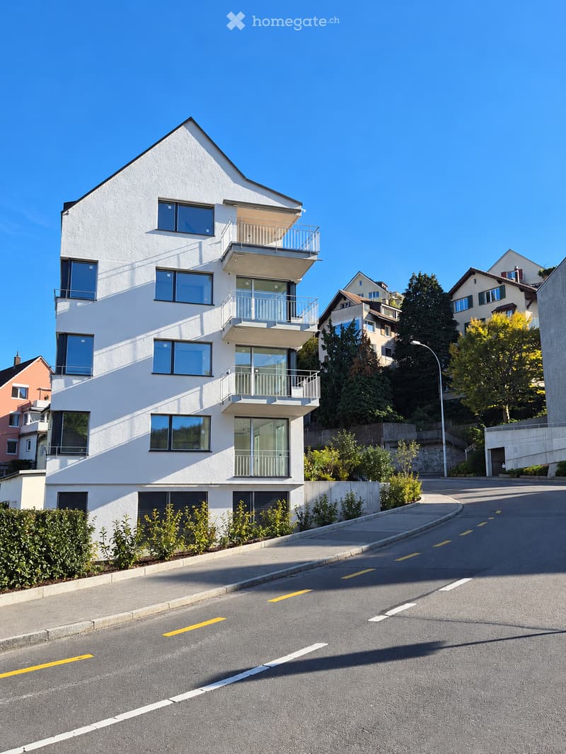 Modern white multi-story building with large windows and balconies, situated on a hillside with neighboring houses and greenery.