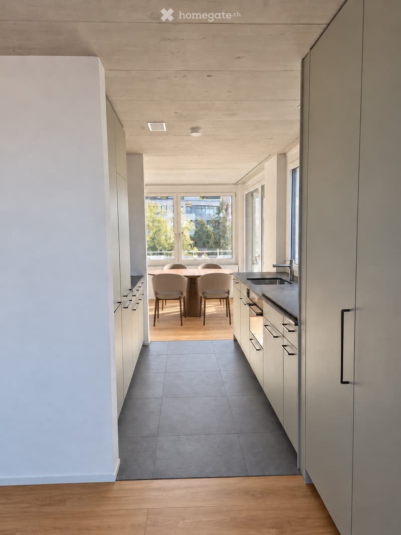 Modern kitchen with white cabinets, dark tiled floor, and a dining area with a round table and four chairs. The space features a sleek sink and faucet, and large windows provide natural light.