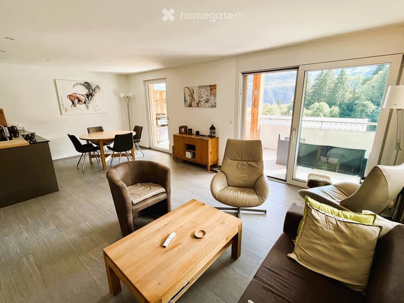 Modern living room with a sofa, coffee table, chairs, dining table, and a view through large windows.