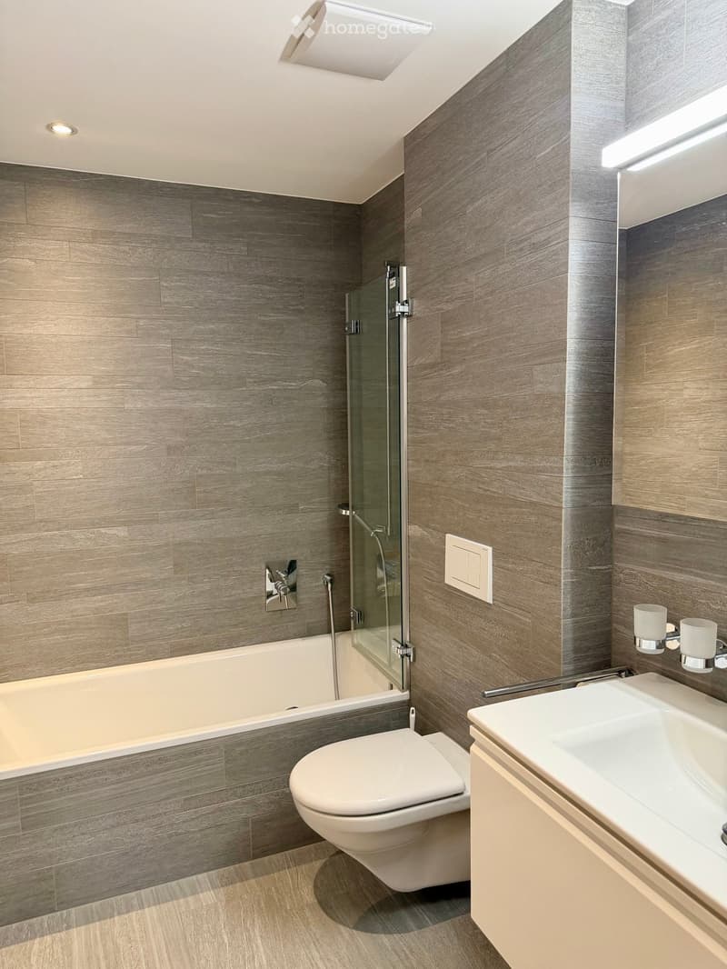 Modern bathroom with grey wood-paneled walls, a bathtub, toilet, sink, and a glass-enclosed shower area.