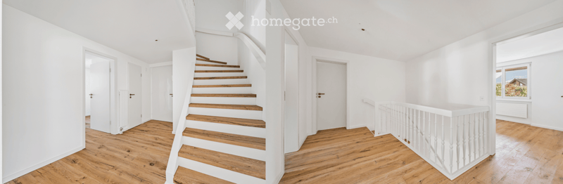 Bright, open hallway with wooden flooring, white walls, and a staircase leading upwards. The space features several white doors and a window on the right side.