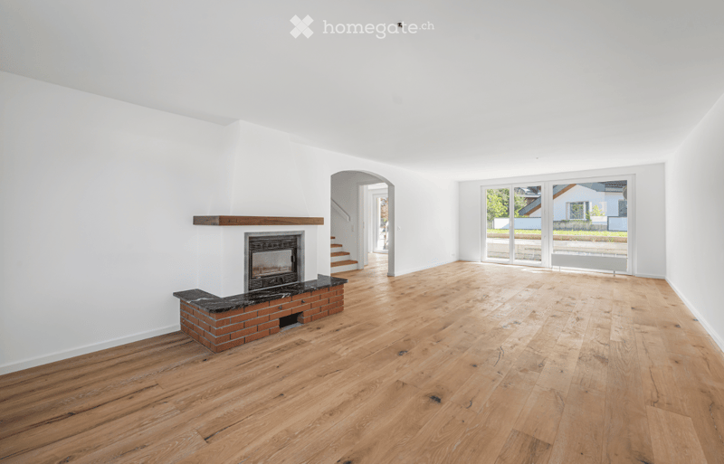 Empty room with wooden flooring, white walls, and a brick fireplace with a black marble mantel. Large windows and an open doorway lead to another space.
