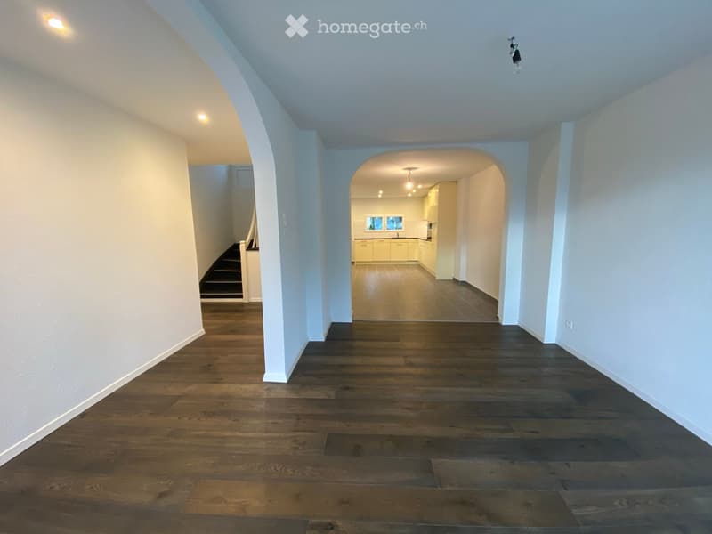 Spacious hallway with wooden flooring, white walls, and arched entryway leading to a kitchen area with light-colored cabinets and stainless steel appliances.