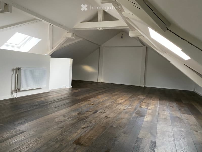 Empty attic space with wooden flooring, white walls, exposed wooden beams, and skylight windows allowing natural light.