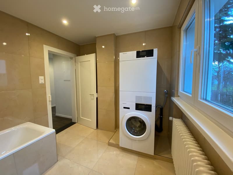 Modern bathroom with a bathtub, a washing machine, and a radiator near the window. The space features beige tiles and a large window offering a view of greenery.