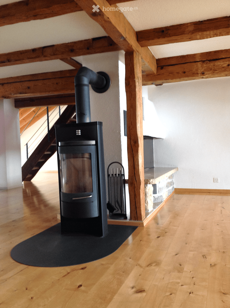 Wooden floor room with a modern wood stove and fireplace tools nearby, exposed wooden beams on the ceiling, and a staircase leading upwards.