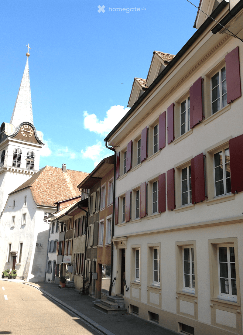 A narrow street lined with traditional European buildings, featuring a prominent church tower with a spire and clock, and colorful shutters on the residential houses.