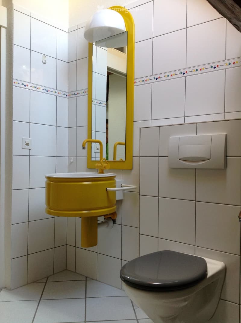 Bright bathroom with white tiled walls and floor, featuring a yellow sink, mirror, and toilet.