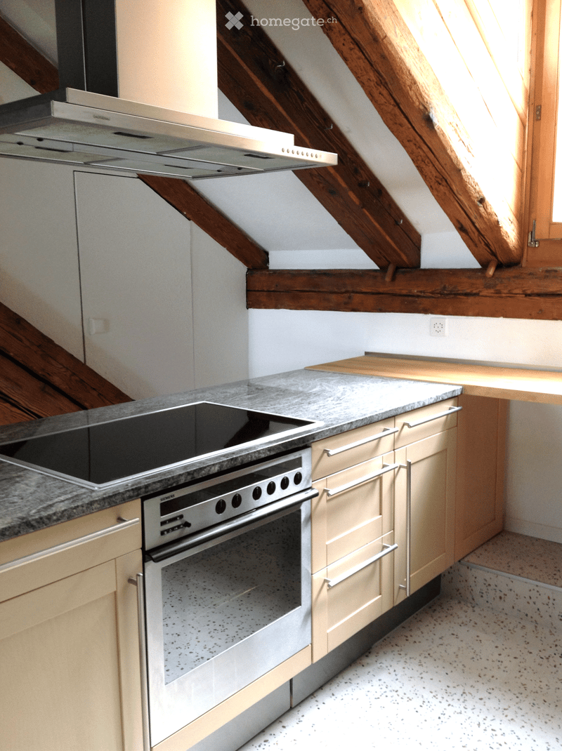 Modern kitchen with light-colored cabinets, granite countertop, built-in oven, and stainless steel range hood. Wooden beams visible in the background.