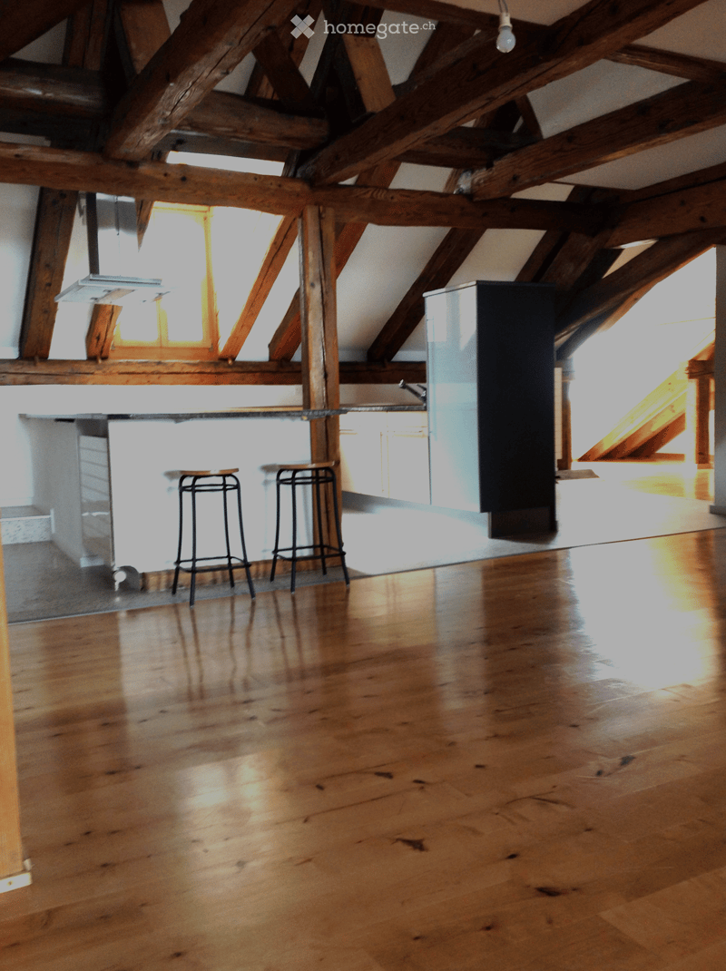 Empty attic space with wooden beams, polished wooden floor, and natural light from skylights. Two bar stools are placed near a counter.