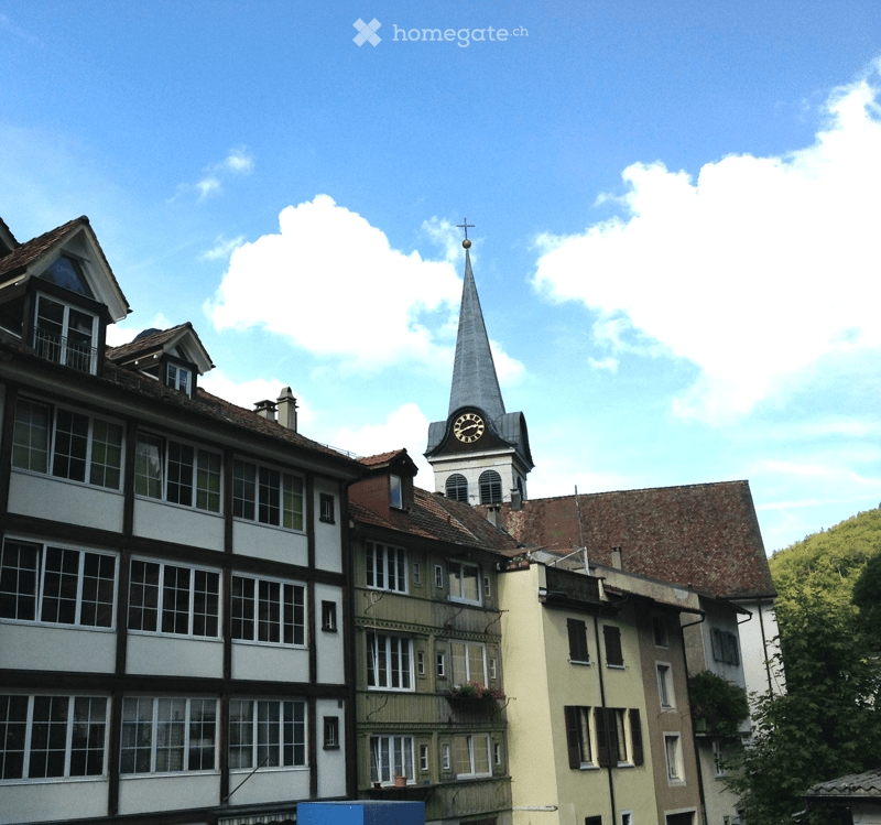 A multi-story building with a prominent clock tower and spire, surrounded by other residential structures with varied facades and windows.