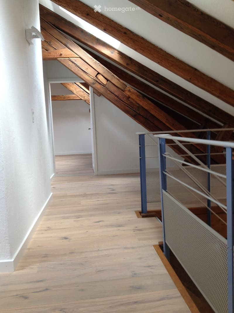 Empty hallway with wooden flooring, white walls, and a staircase with metal railings leading to an attic space with exposed wooden beams and skylights.