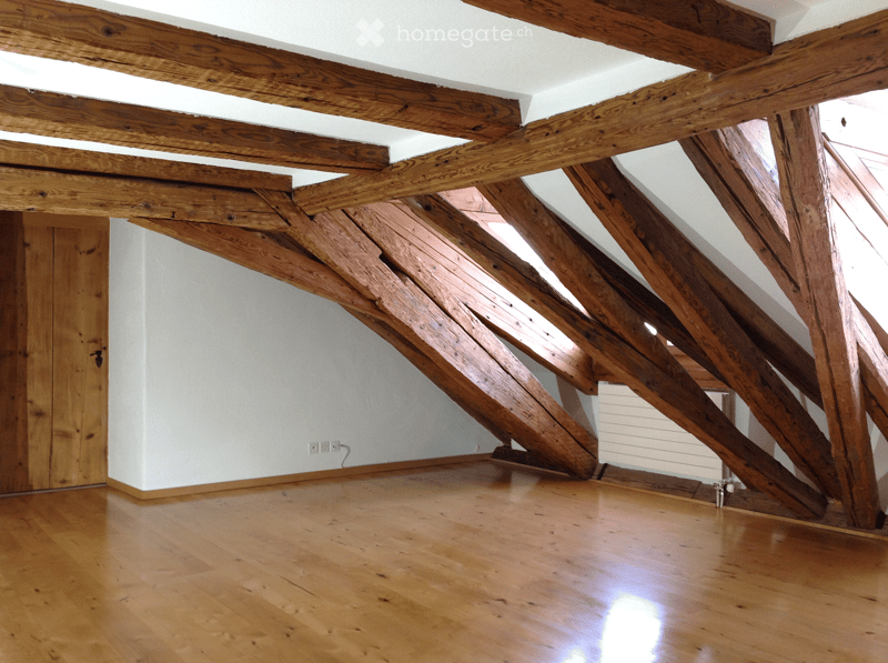 Empty attic space with exposed wooden beams, wooden floor, white walls, and a small wooden door.