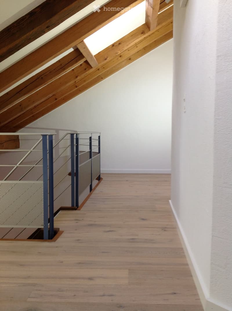 Empty room with wooden floor and exposed wooden beams on the ceiling. A metal staircase with a railing is visible on the left side.