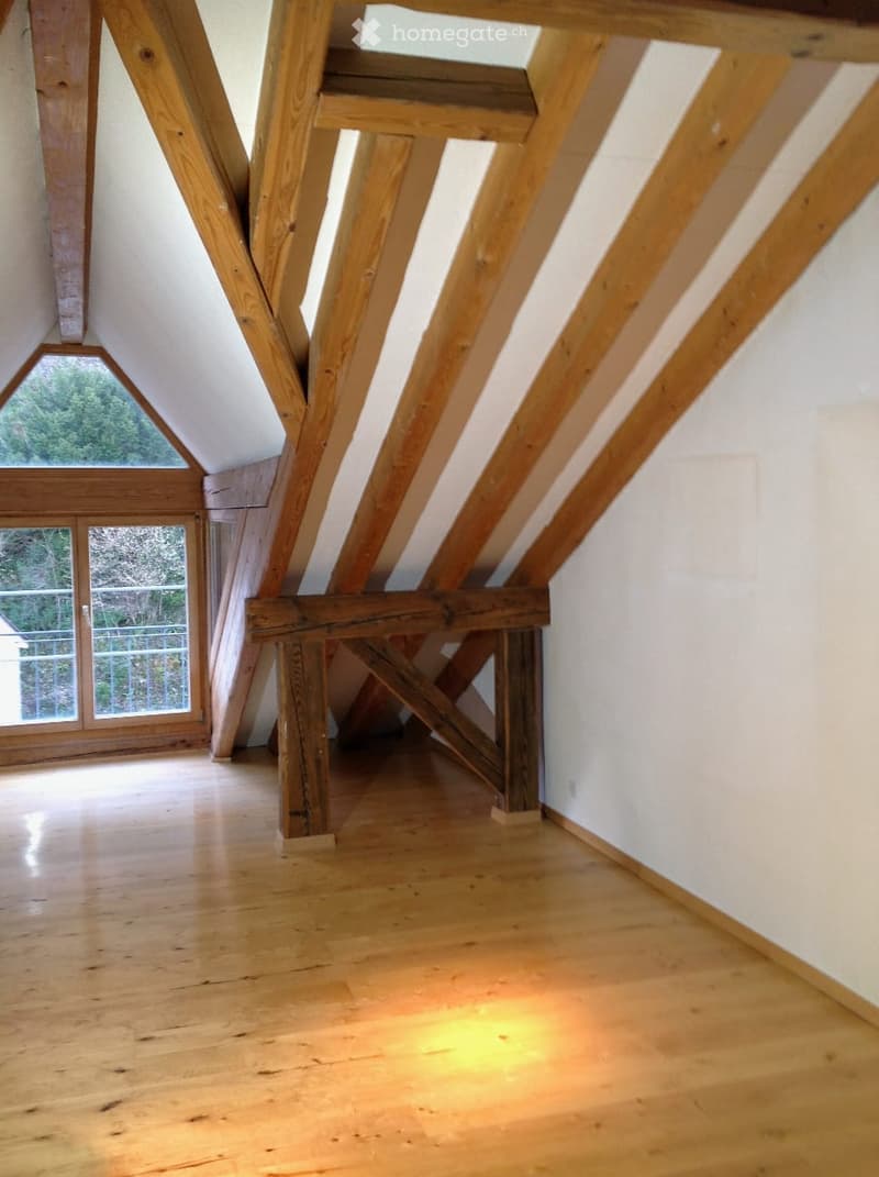 An empty attic space with exposed wooden beams, wooden flooring, and large windows offering a view of greenery outside.