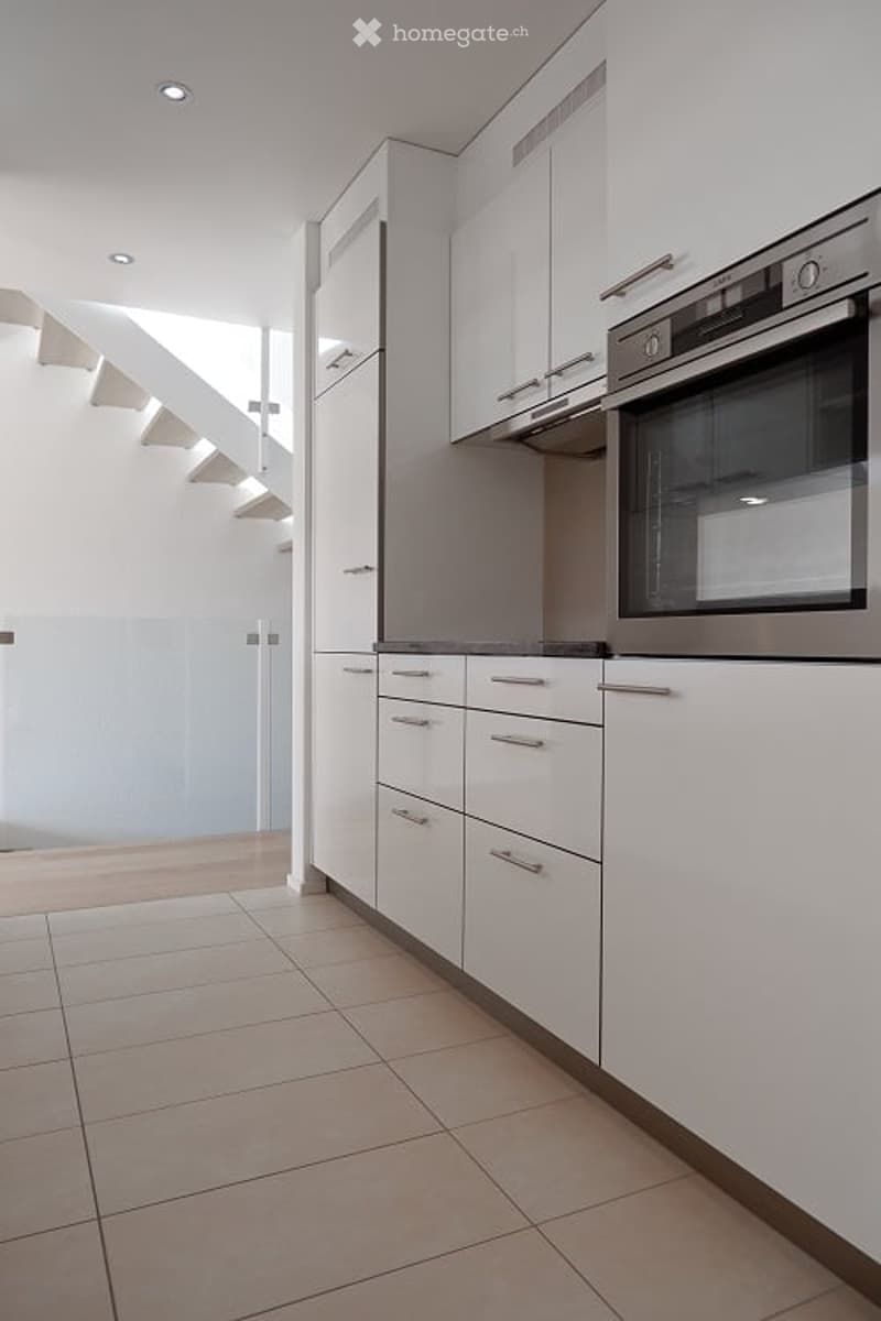 Modern kitchen with white cabinets, tiled floor, stainless steel oven and dishwasher, and a staircase in the background