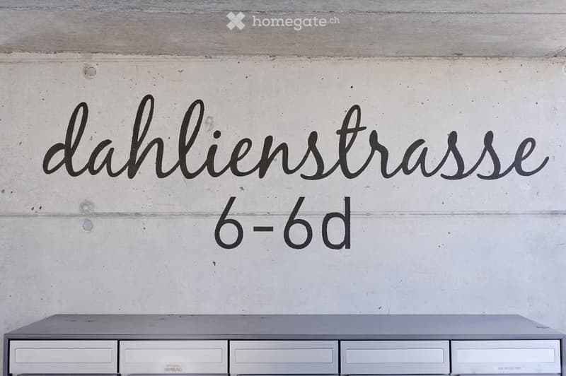 A modern, minimalist room with a sleek grey cabinet featuring multiple drawers and a smooth surface, set against a concrete wall with a decorative address sign reading "dahlienstrasse 6-6d".