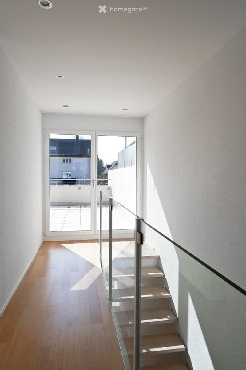 A corridor with wooden flooring, a sliding glass door, and a staircase with a glass railing. The wall is white with recessed lights, and the balcony has railings. The floor is shiny, and the room is bright.