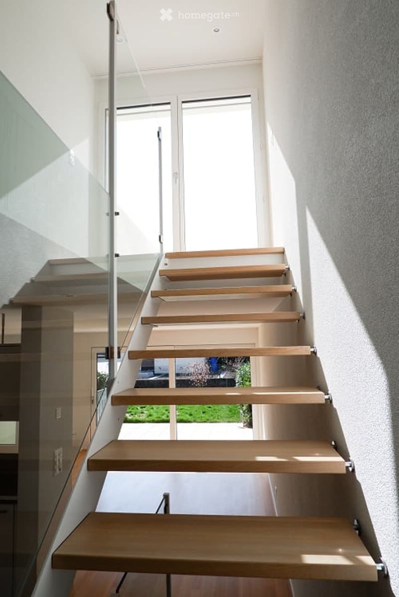 The house has a modern staircase with wooden steps and glass railings, leading to an upper floor. The staircase is placed against the wall, with white walls and a clear view outside the windows. The staircase has a simple design with no additional feature