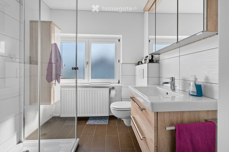 Modern bathroom with a white sink, chrome faucet, mirror, toilet, and a glass-enclosed shower. Wooden cabinets and a purple towel are visible.