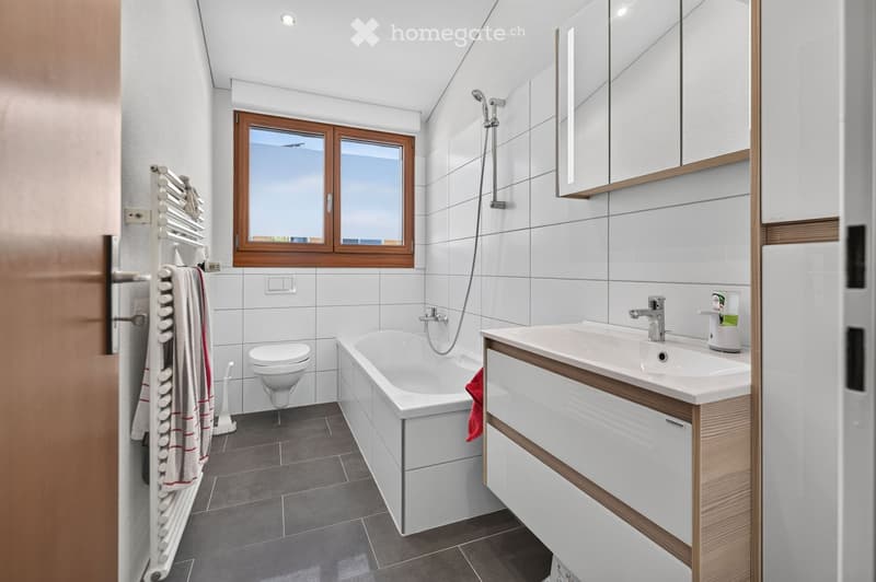 Modern bathroom with white tiles, a bathtub, sink, toilet, mirror, towel rack, and a window with wooden frame.