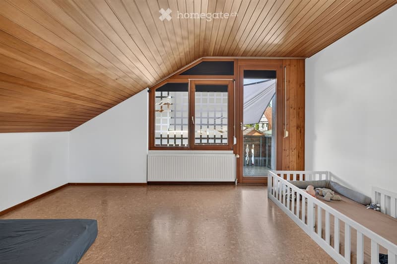 Bright, airy room with wooden ceiling, large windows, and a crib with toys. The space includes a radiator and a sliding glass door leading outside.