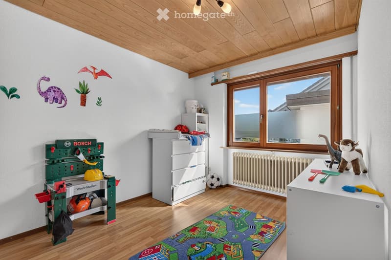 Brightly lit children's room with wooden flooring, a toy workbench, storage unit, play mat, and various toys including a dinosaur and animal figures.