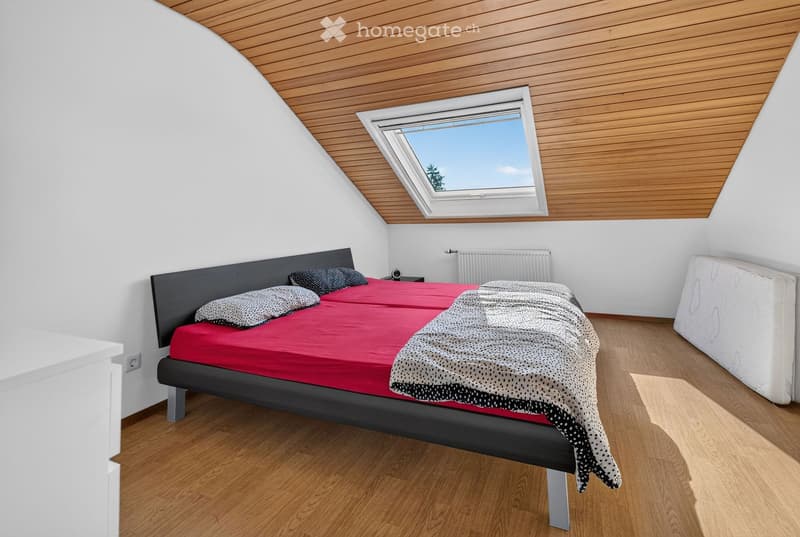 Modern bedroom with wooden flooring, slanted wooden ceiling, skylight window, double bed with red bedding and patterned pillows, nightstand, and radiator.